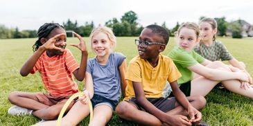 Five children sitting on grass, smiling and chatting during outdoor playtime.