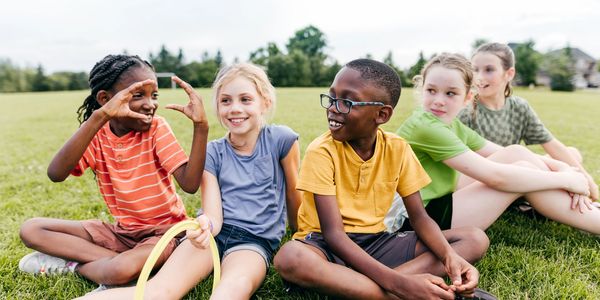 Five children sitting on grass, smiling and chatting during outdoor playtime.