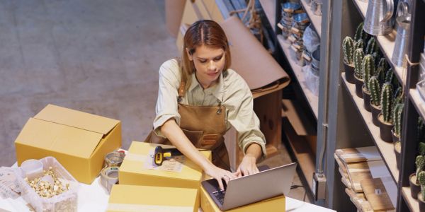 Woman working on laptop amid packaging boxes and plants.