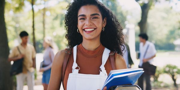 Smiling student holding books outdoors on a sunny day.