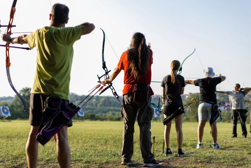 Group of people practicing archery on the training field . They are standing in a row, holding bows and aiming at target.