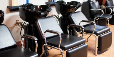 Black salon chairs lined up in a hair salon beside wash basins.
