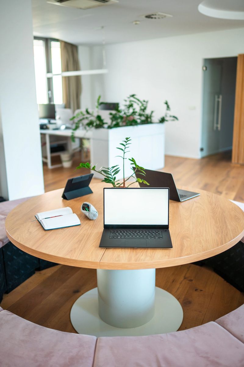 A sleek laptop, smartphone, and notepad rest on a round wooden desk in a chic office, reflecting a professional's tools in a dynamic work environment.