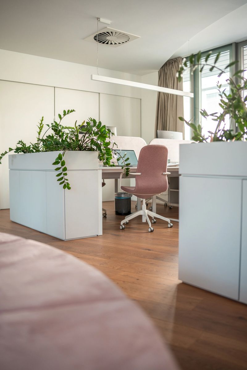 A spacious office with sleek white desks adorned with green plants, comfortable rolling chairs, and multiple tech devices under bright lights.