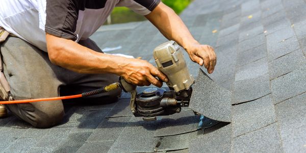 Worker installing gray asphalt shingles on a roof with a nail gun.