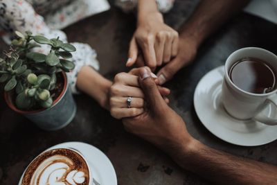 Couple holding hands with an engagement ring, coffee cups, and a plant on the table.