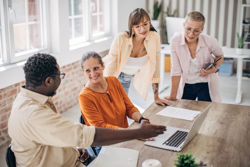 A group of business professionals engaging in an active brainstorming session around a laptop in a bright, modern meeting room, emphasizing collaboration and creativity.