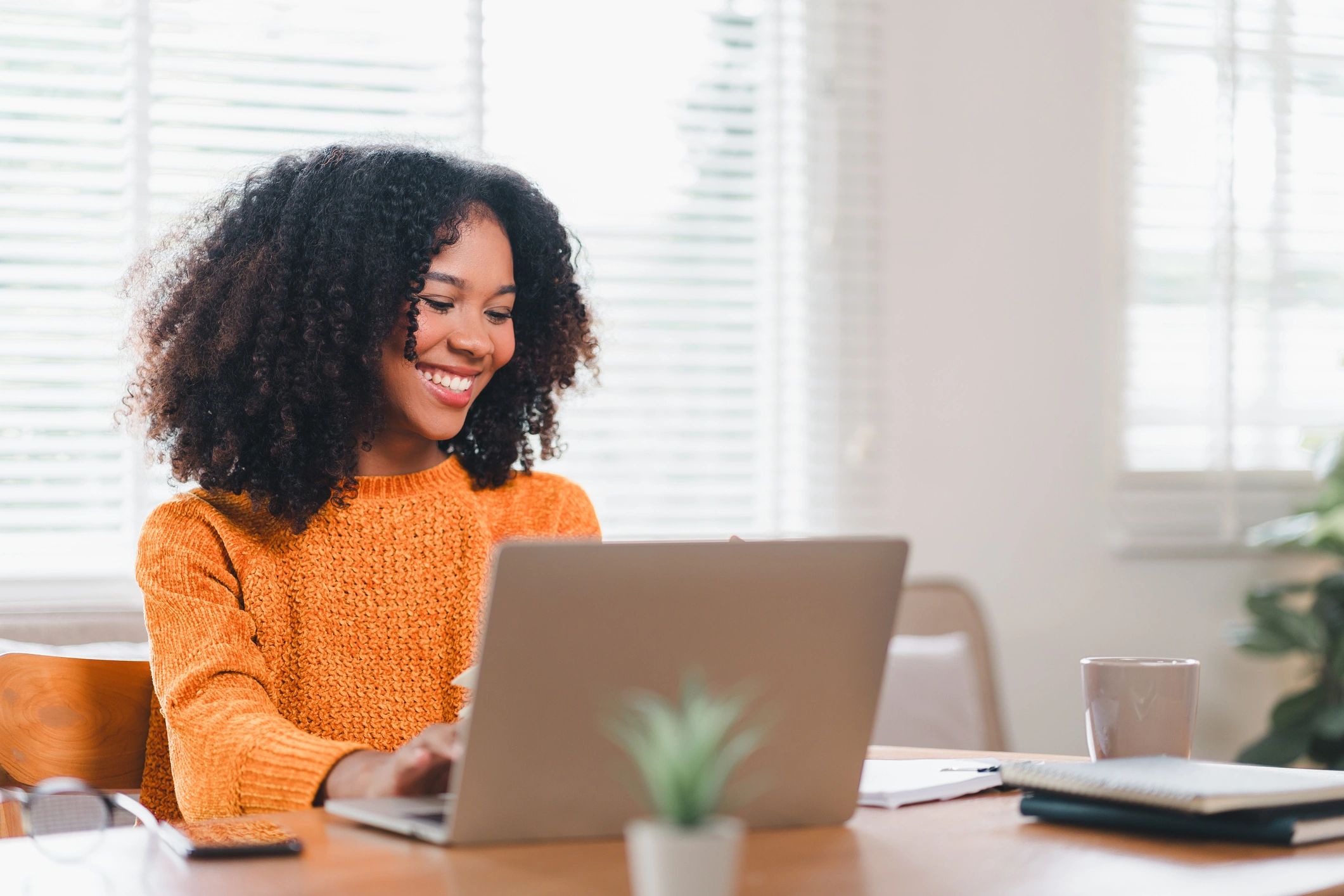 Smiling woman in orange sweater working on a laptop at a bright table.