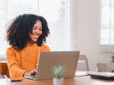 Smiling woman in orange sweater working on a laptop at a bright table.
