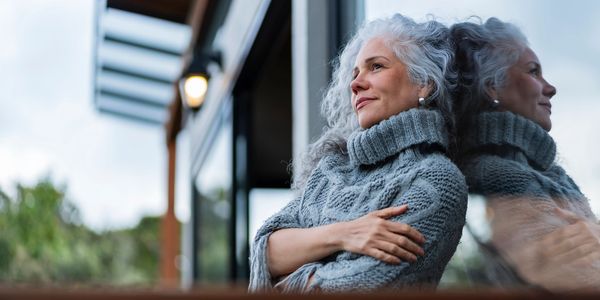 A woman in a cozy sweater gazes thoughtfully outside, reflected in a glass window.