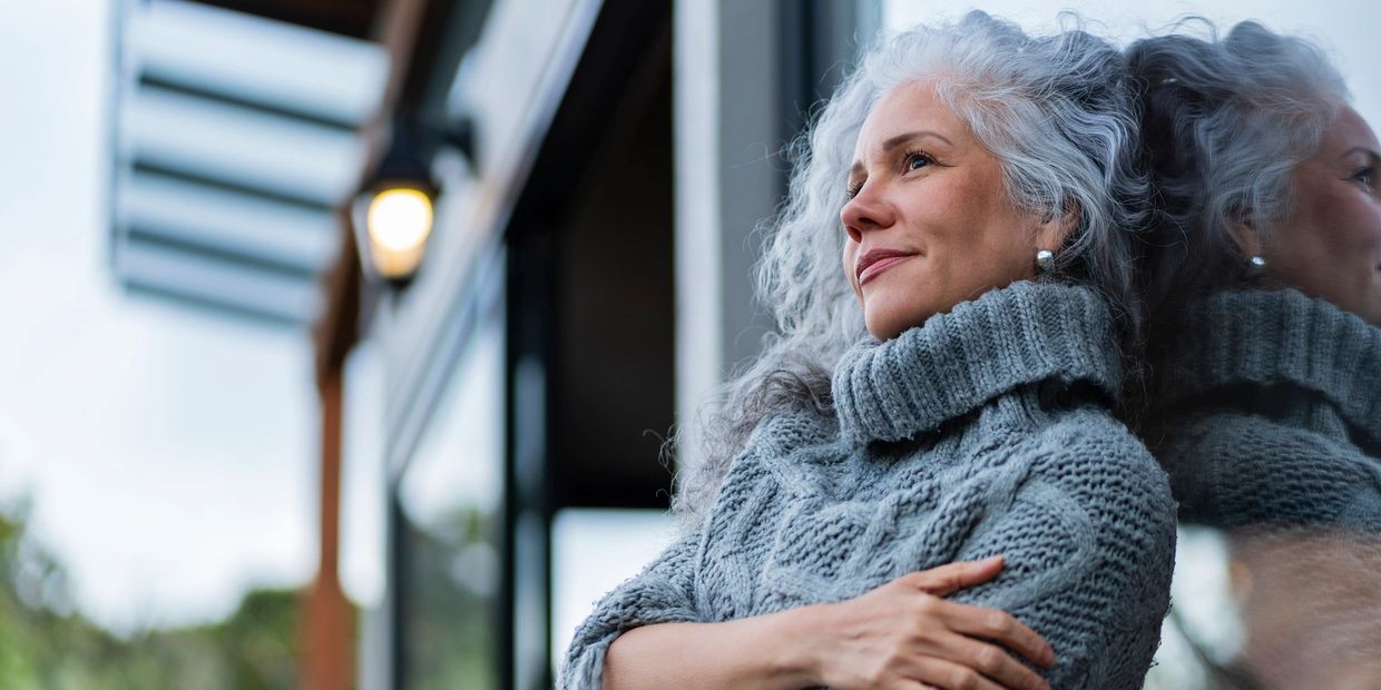 A woman in a cozy sweater gazes thoughtfully outside, reflected in a glass window.