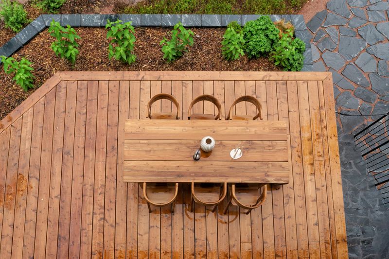 A stunning wooden terrace table group, seen from top to bottom. Terrace is beautifully bordered with rain-refreshed garden plants.