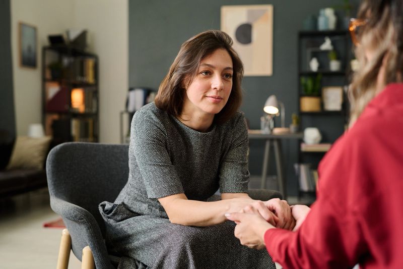 Female therapist sitting opposite to client, holding hands in supportive gesture during counseling session in well-decorated office environment