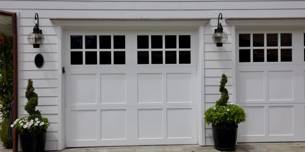 Two white paneled garage doors with black window panes and decorative plants.