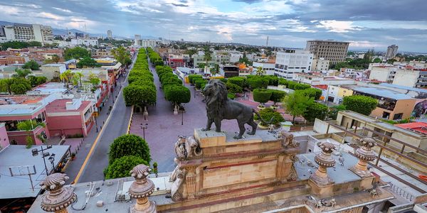 Historic arch with a lion statue overlooking a tree-lined boulevard in a vibrant city.