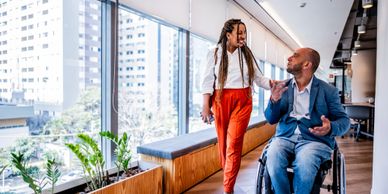 A woman and a man in a wheelchair share a joyful moment in an office corridor.