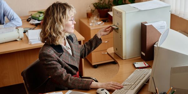 A woman inserts a floppy disk into a vintage computer at her office desk.