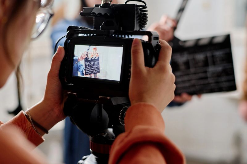 Close-up view of camera display showing clapperboard during film shoot. Person holding camera with crew member clapping in background