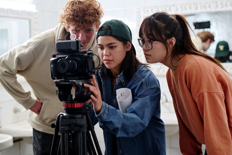 Group of young diverse individuals working together while setting up professional camera in modern studio environment. Enthusiastic team reviewing footage on camera screen