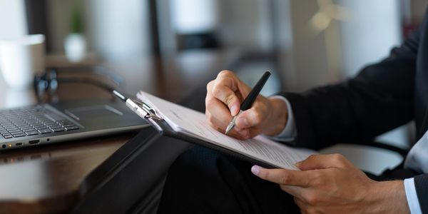 Person signing a document on a clipboard at a desk with a laptop nearby.