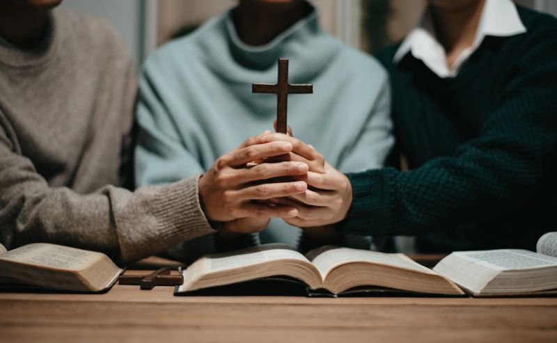 Group of Christians sit together and pray around a wooden table with blurred open Bible pages in their homeroom. Prayer for brothers, faith, hope, and seek the blessings of God.