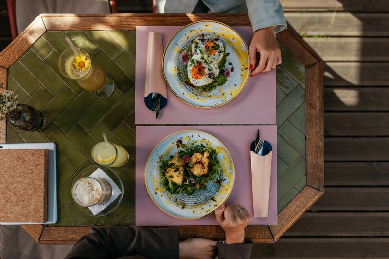 Top view of a table on a wooden floor at a cafe with breakfast food and drinks on it and two people sitting at the table.