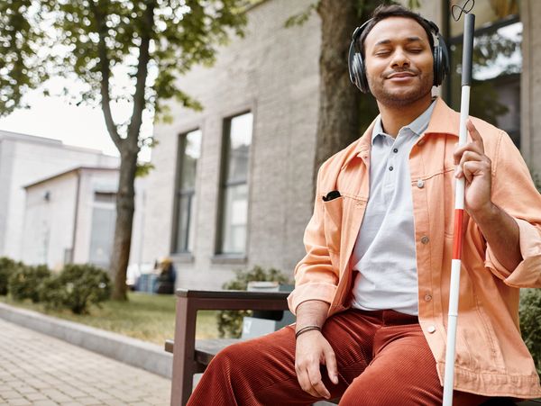 A man sitting on a bench with his white cane wearing headphones