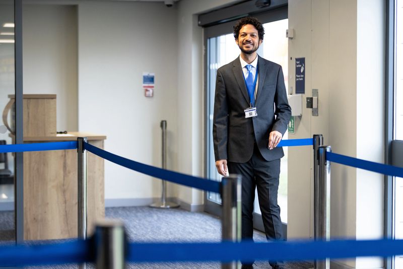 Wide view of a mid adult male attendant waiting for customers to arrive at the check in desk so he can check boarding passes and safely allow passengers onto the flight. He is wearing a suit and tie with a lanyard to display his staff ID.