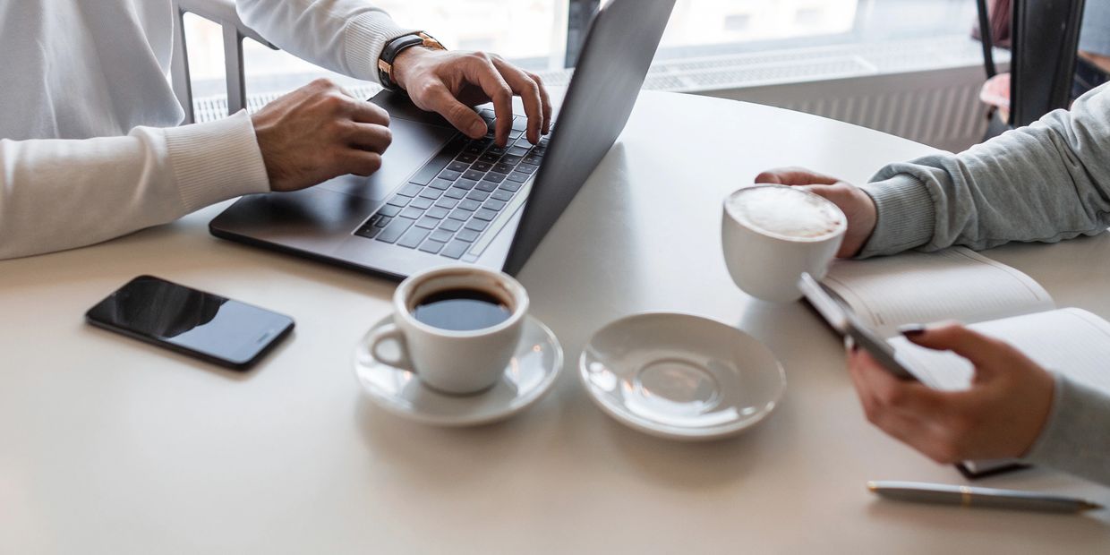 Two people working with coffee, laptop, and phone at a table by a window.