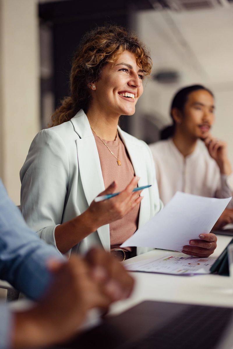 Confident businesswoman smiles during an office meeting, holding papers and engaging with colleagues in a professional environment.