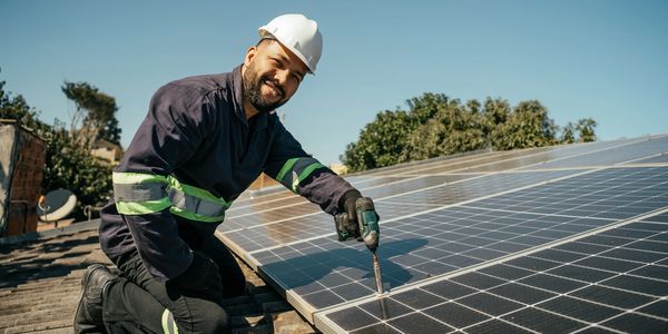 A worker installs solar panels on a roof under clear skies.