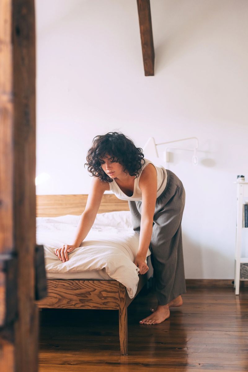 A young woman with curly hair is tidying up a bed in a warm, sunlit bedroom. The atmosphere is peaceful and serene, conveying a sense of comfort and home.