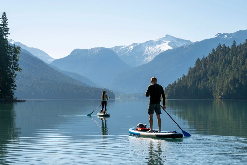 Couple paddle SUP boards across mountain lake, Cheakamus Lake, Whistler