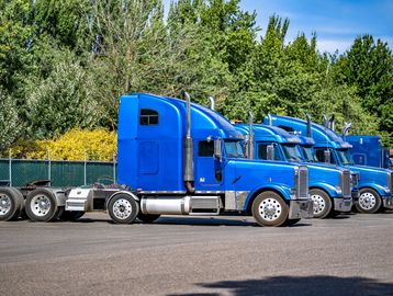 Row of blue semi trucks parked in a lot with trees in the background.