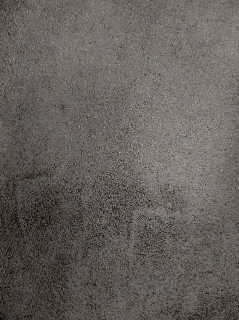 A close-up image of a weathered concrete wall with a rough, uneven texture. The surface is a muted gray color with darker patches and lighter streaks. The texture is reminiscent of fine grains of sand or small pebbles embedded in the concrete.