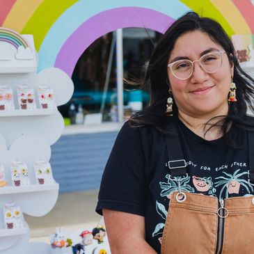 Smiling woman wearing glasses and colorful earrings stands by a rainbow-themed display of cute accessories.