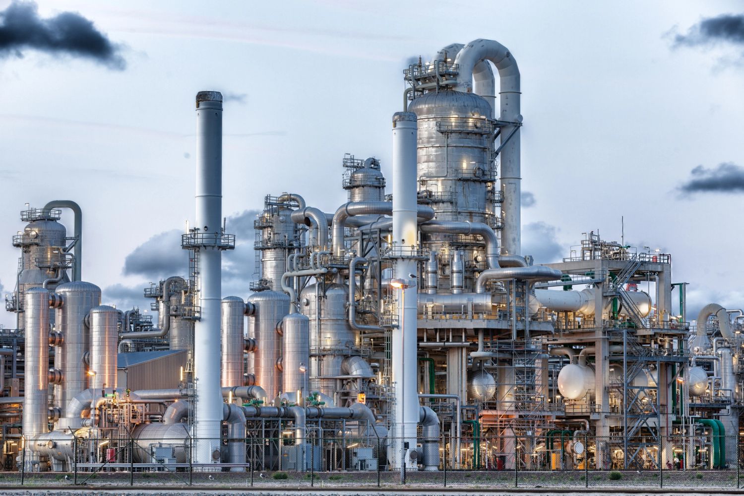 Large industrial refinery with numerous silver pipes and chimneys emitting black smoke.