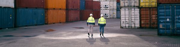 Two workers walk between rows of stacked shipping containers at a port.