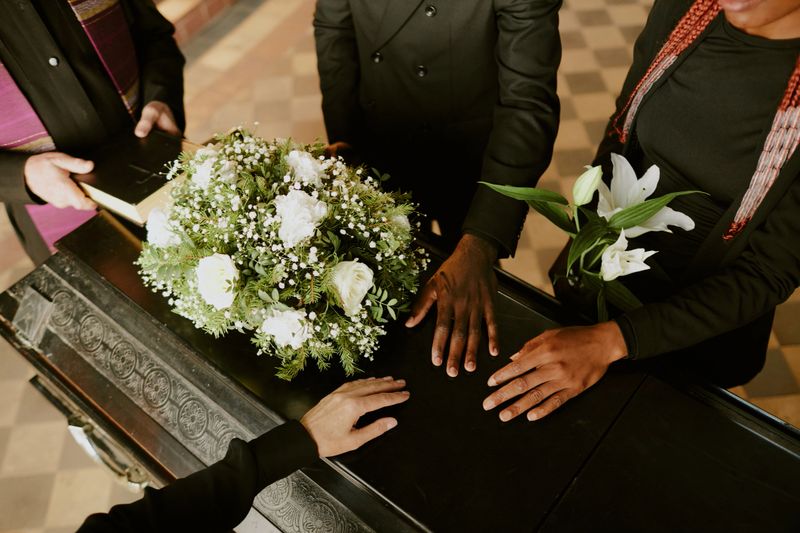 High angle shot of unrecognizable hands on black wooden coffin with white roses on top, pastor holding holy book