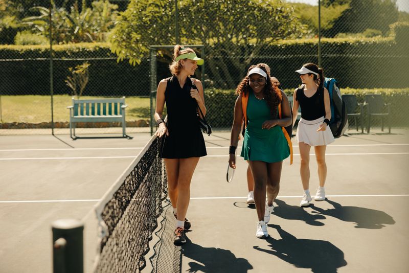 Three women tennis players in sportswear, carrying sports bags, walking together on a court near the net and bench after a game.