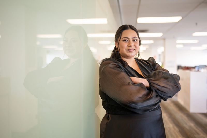 A confident Māori woman stands with arms crossed in a sleek, contemporary office setting. Her stylish attire reflects her sense of empowerment and professionalism