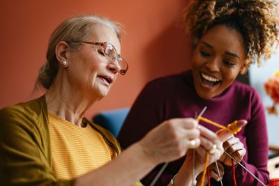 Two women joyfully knitting together indoors.