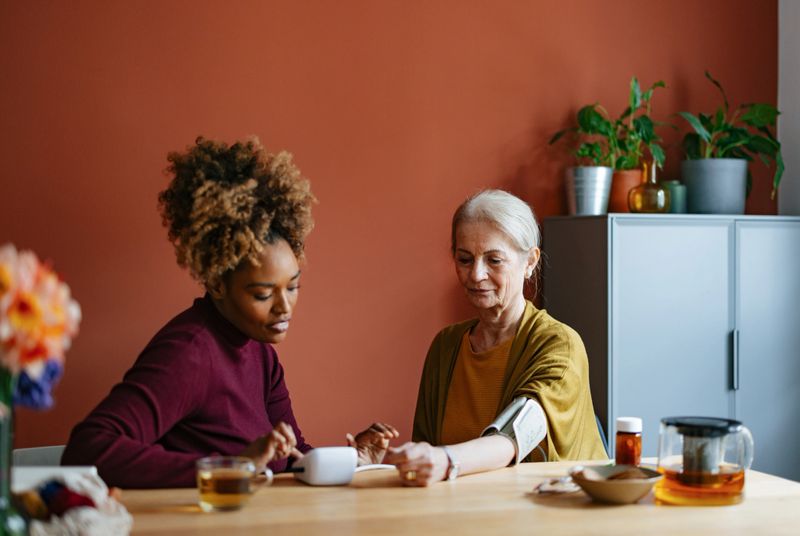 A caregiver helps an elderly woman monitor her blood pressure at home. The scene depicts care, trust, and companionship in a warm, comfortable setting.