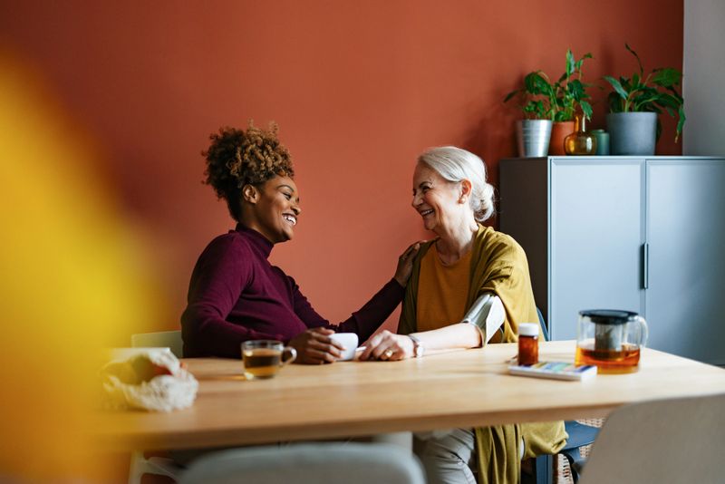 Senior woman sharing a joyful moment with her caregiver at a cozy home setting. They are sitting at a table, smiling and enjoying tea. This image captures warmth, companionship, and care.