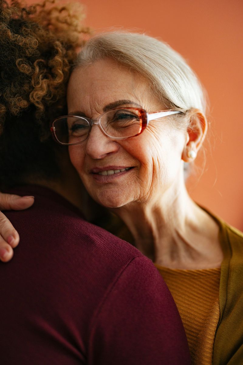 An elderly woman wearing glasses shares a warm embrace with a caregiver or family member, conveying love, support, and companionship in a cozy setting.
