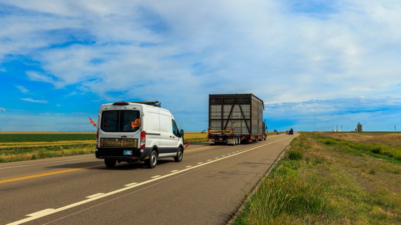 Kansas, USA - May 31. 2024:Oversized load convoy travels along a rural Kansas highway. Large truck carries a massive metal structure, escorted by a white van. Road stretches through flat farmland under partly cloudy sky with cirrus cloud streaks. Utility poles line roadside, grain silo visible in distance