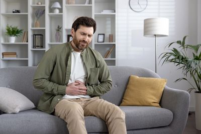 A man sitting on a couch, clutching his stomach in discomfort.