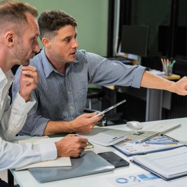 Two men discussing data at a desk with documents and a tablet.
