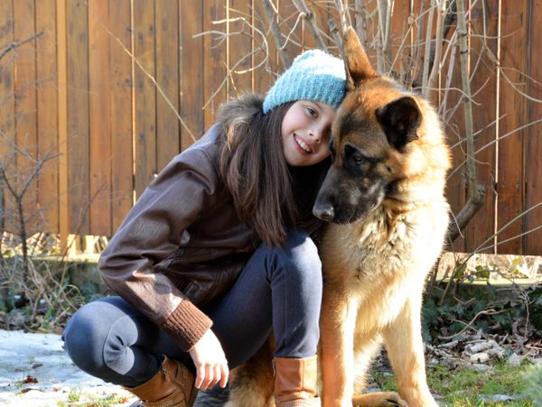 A girl in a blue hat cuddles her large German Shepherd dog outdoors.