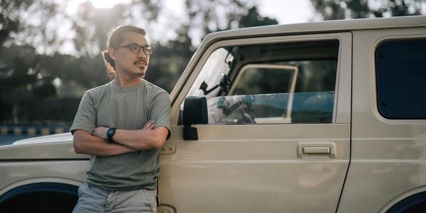 Man with glasses leans on beige SUV with arms crossed, looking to the side.